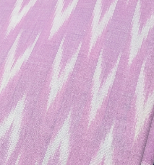 A hand holding a folded pink fabric with a white ikat pattern, with other patterned fabrics stacked underneath.