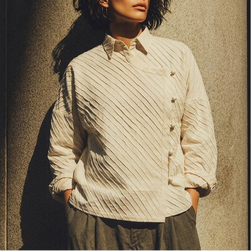 A woman wearing a cream-colored, vertically textured button-up shirt and dark gray trousers, standing against a textured concrete wall in warm, directional sunlight.