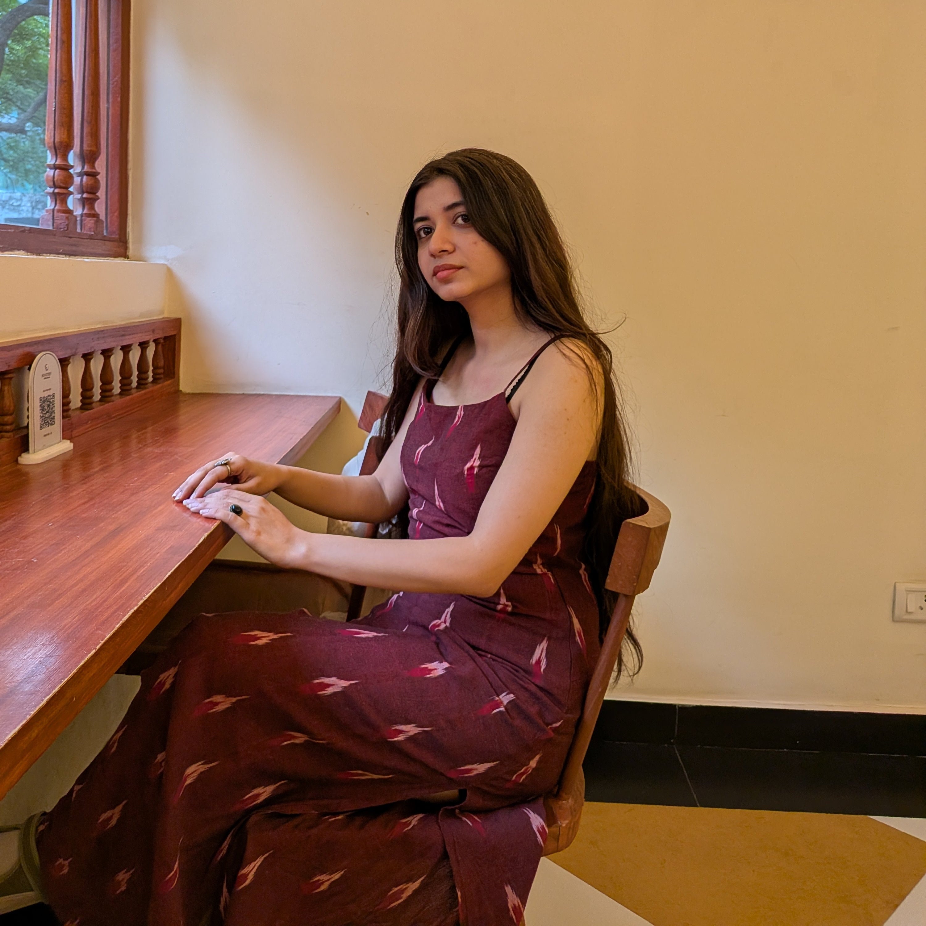 A woman with long dark hair sits at a wooden counter in a cafe, wearing a sleeveless maroon dress with a small pink and white pattern.