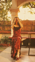 A woman sitting outdoors on a bench in a warm, vintage-style photograph. She is wearing a dark red and black patterned maxi dress with a sheer black strapless overlay and dark pointed-toe heels.